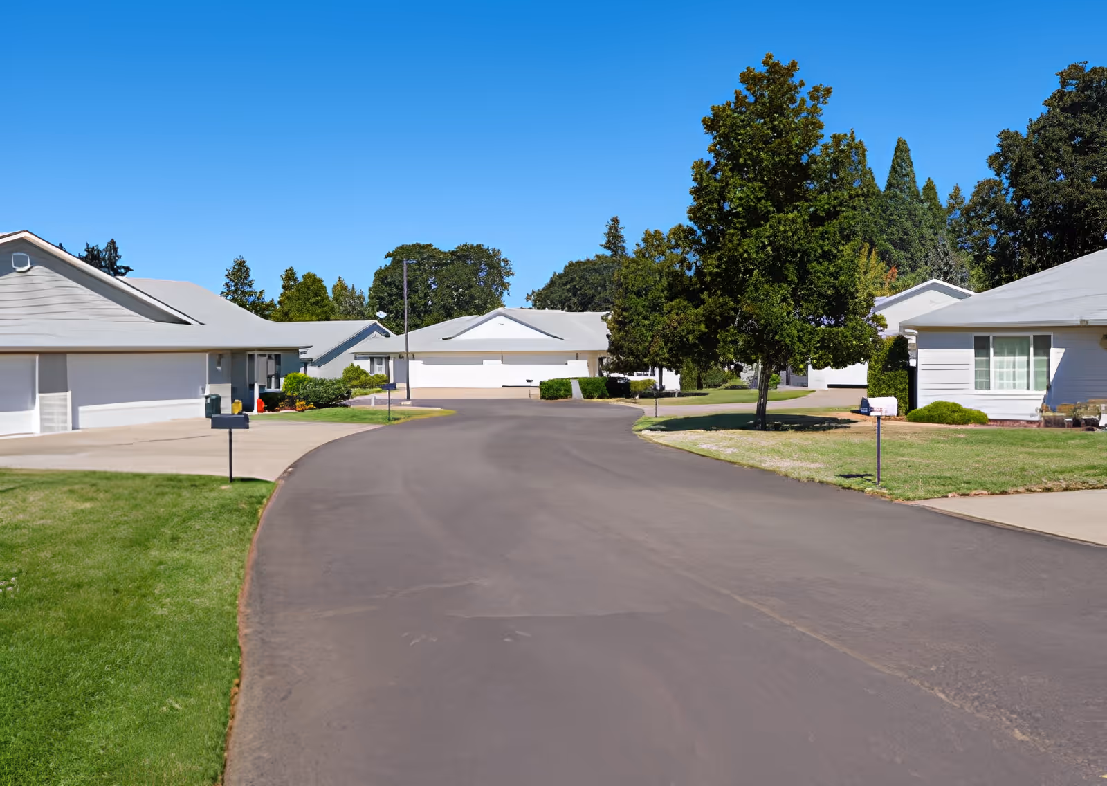 Curved residential street lined with single-story white houses, lawns, and trees under a clear blue sky.