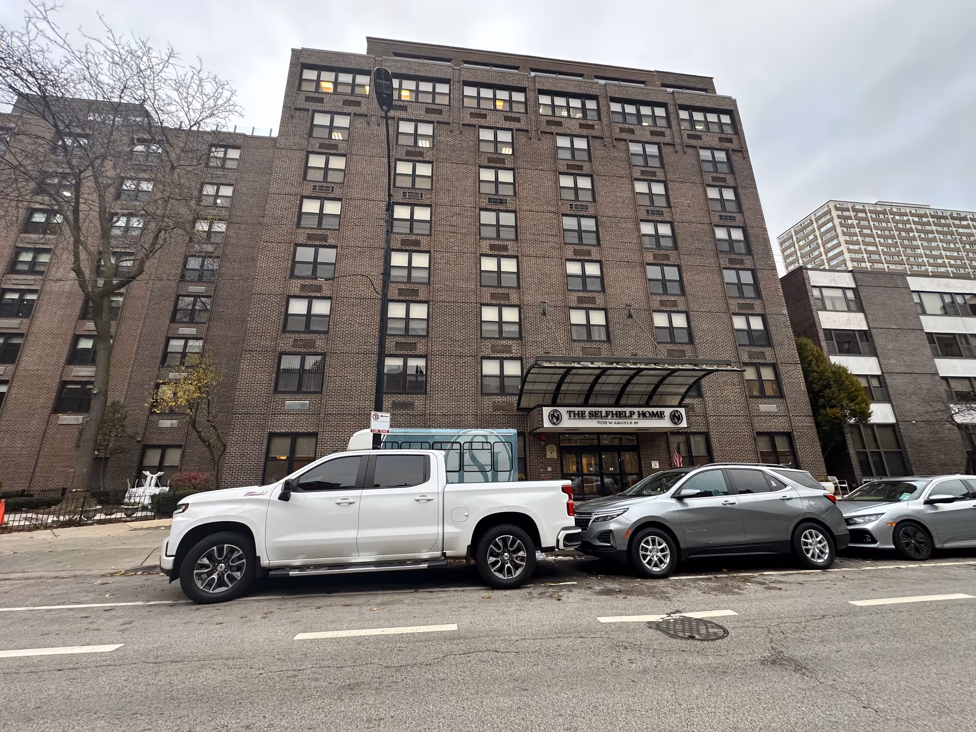 Exterior view of The Selfhelp Home Senior Living Community building, a multi-story brick structure with many windows. Several cars are parked along the street in front of the building. The entrance has a canopy with the facility's name displayed above the doors.