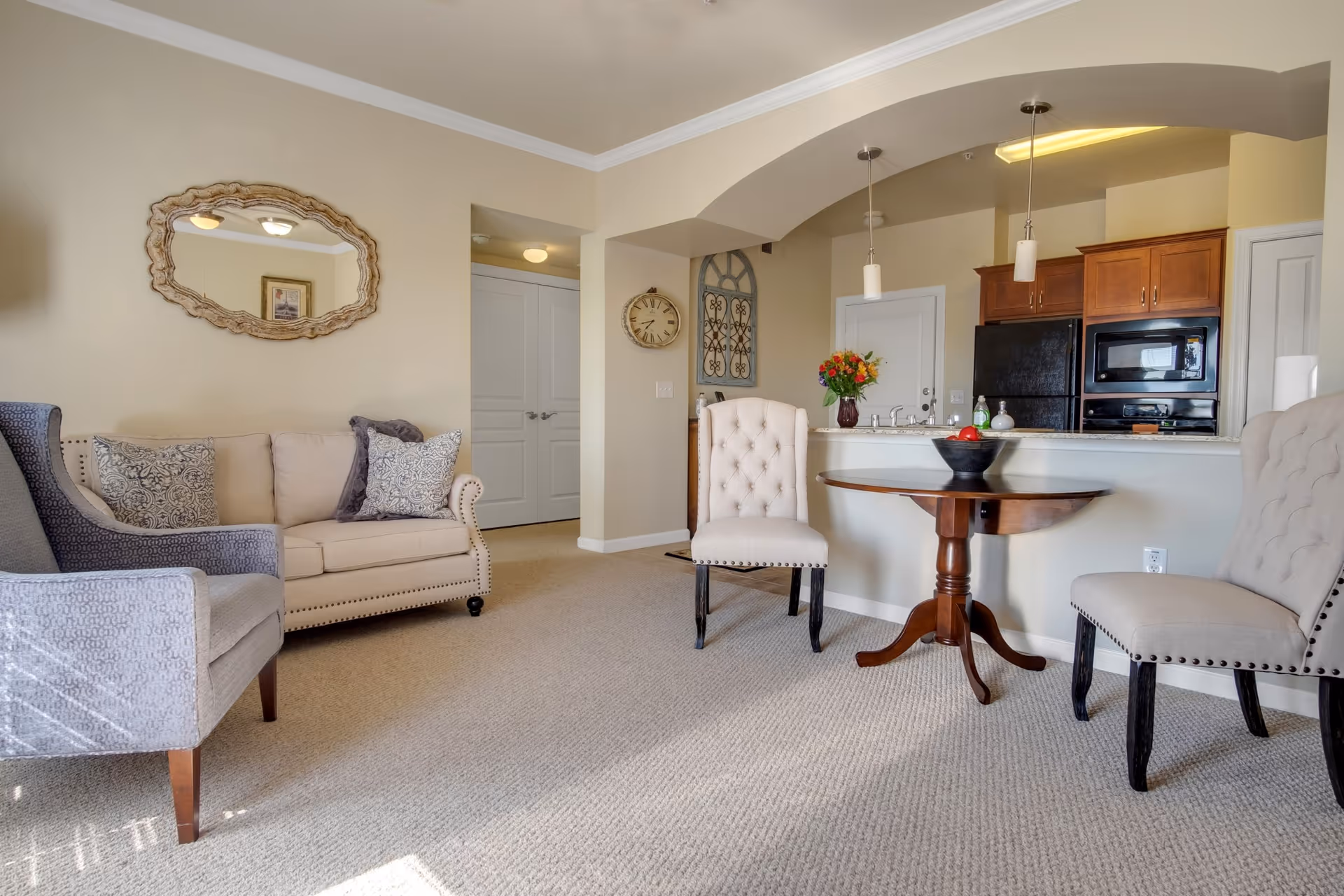 A cozy living room area with beige walls and carpet. The room features a beige loveseat with patterned pillows, a gray armchair, and two beige upholstered chairs around a small wooden round table with a bowl of fruit. In the background, there is a kitchen with wooden cabinets, a black refrigerator, and a microwave. Decorative elements include a wall mirror, a clock, and a floral arrangement on the kitchen counter.
