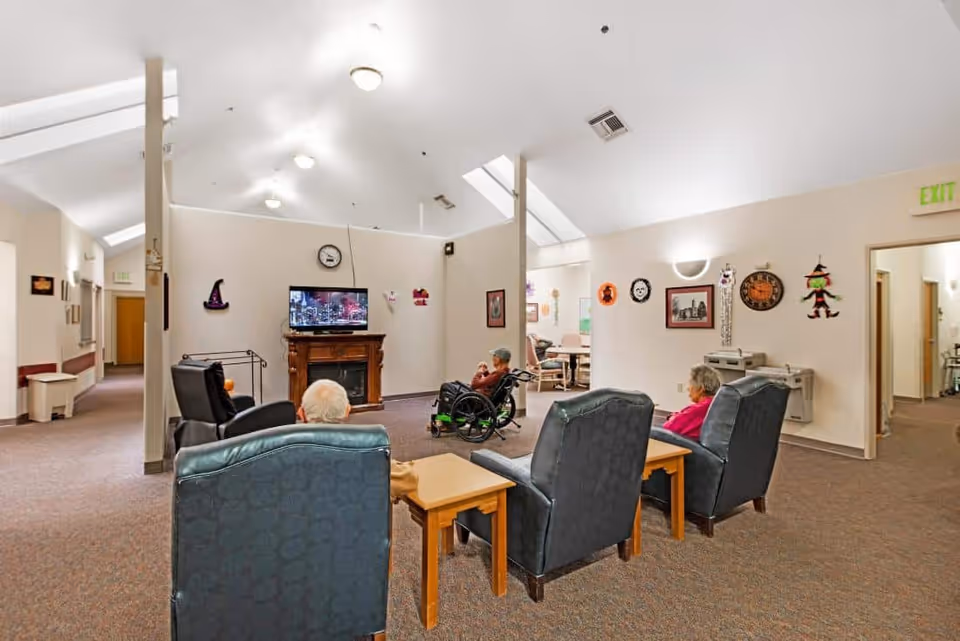 A common area in a senior living facility with several elderly residents seated in armchairs and a wheelchair, watching a television mounted above a wooden fireplace. The room has beige walls decorated with Halloween-themed decorations and framed pictures. The ceiling is high with skylights and ceiling lights, and there is an exit sign above a doorway leading to another room.