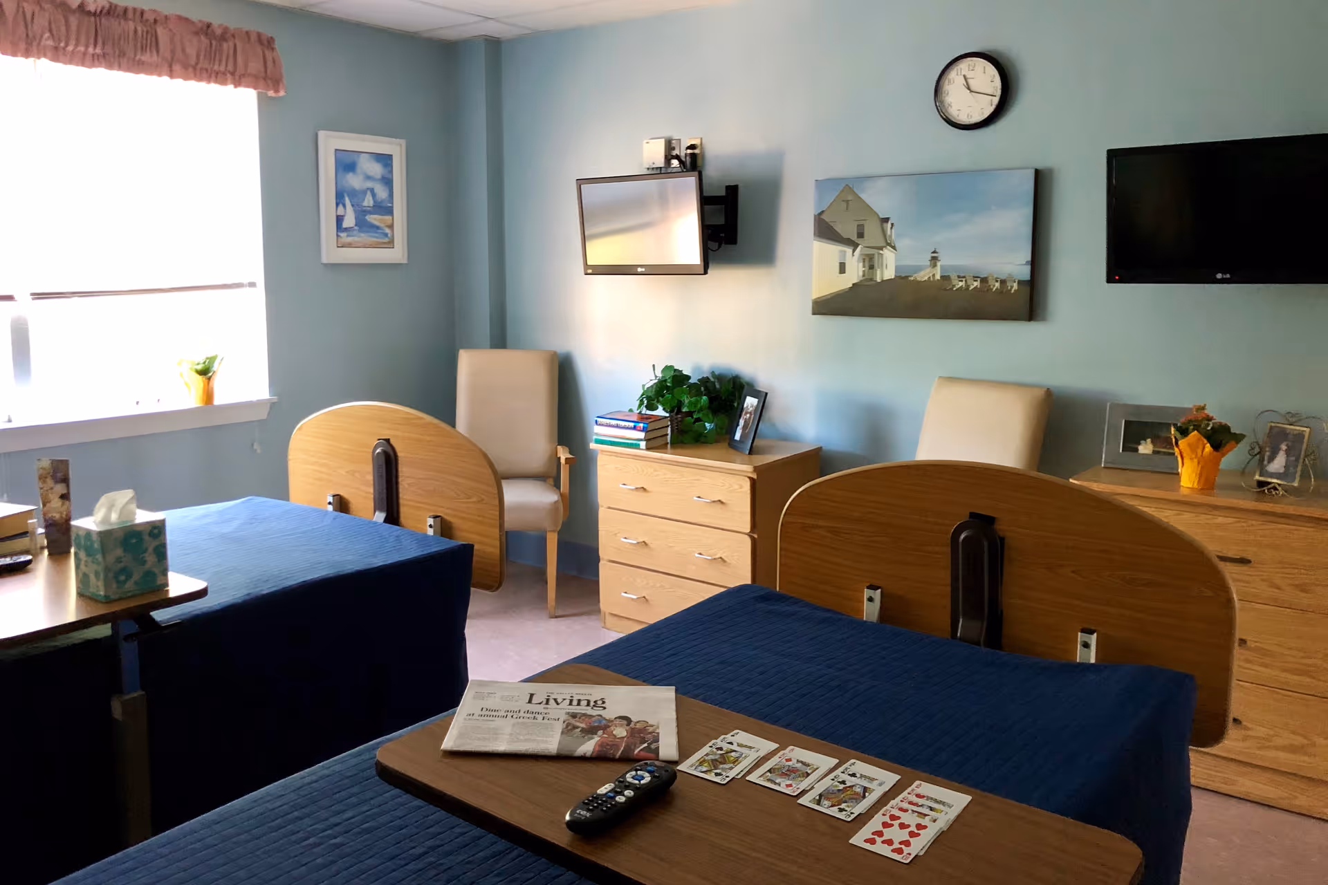 A room with two hospital-style beds covered with blue blankets, each with a wooden headboard. A small table is placed over one bed with a newspaper, playing cards, and a TV remote on it. The room has light blue walls with two paintings, a clock, two beige chairs, two wooden dressers with plants and framed photos, and two wall-mounted flat-screen TVs.