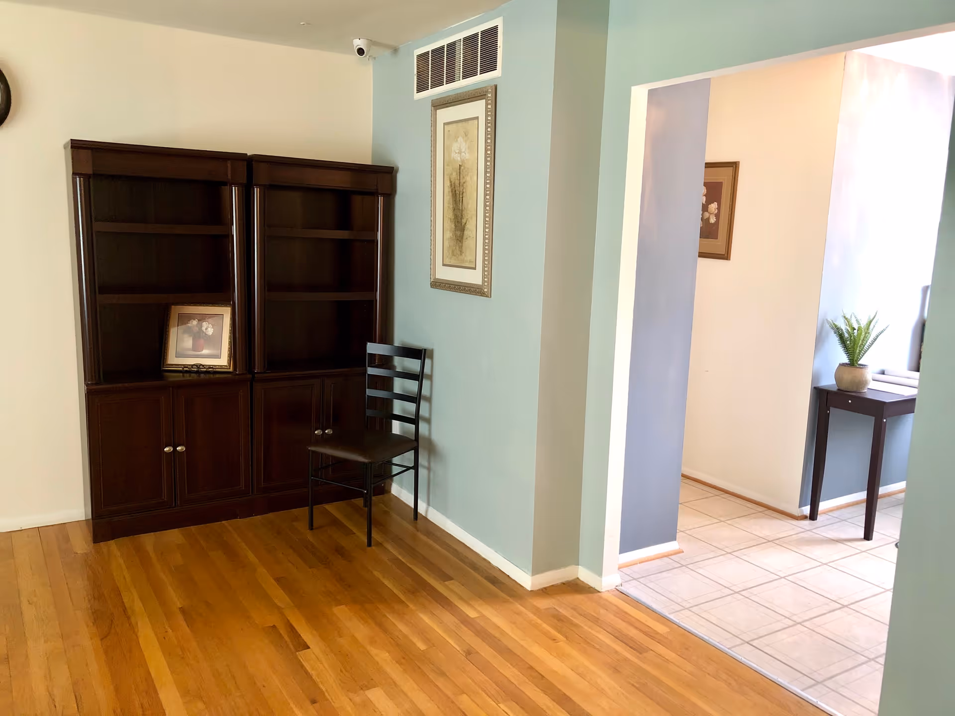 An interior room with wooden flooring featuring two dark wooden bookshelves, a black metal chair with a brown seat cushion, and a framed floral picture on the wall. Adjacent to this room is a tiled floor area with a small dark wooden table holding a potted plant and another framed picture on the wall.