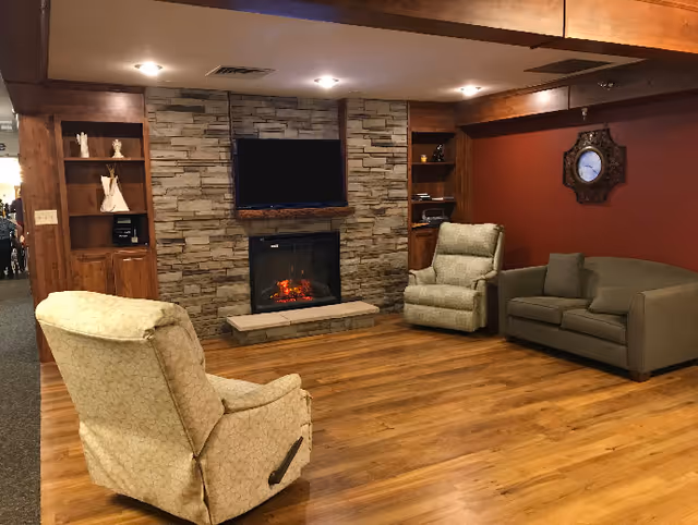 A cozy living room area with wooden flooring, two beige recliner chairs, a gray sofa, a stone fireplace with a mounted flat-screen TV above it, built-in wooden shelves on either side of the fireplace, and a decorative wall clock on a red accent wall.