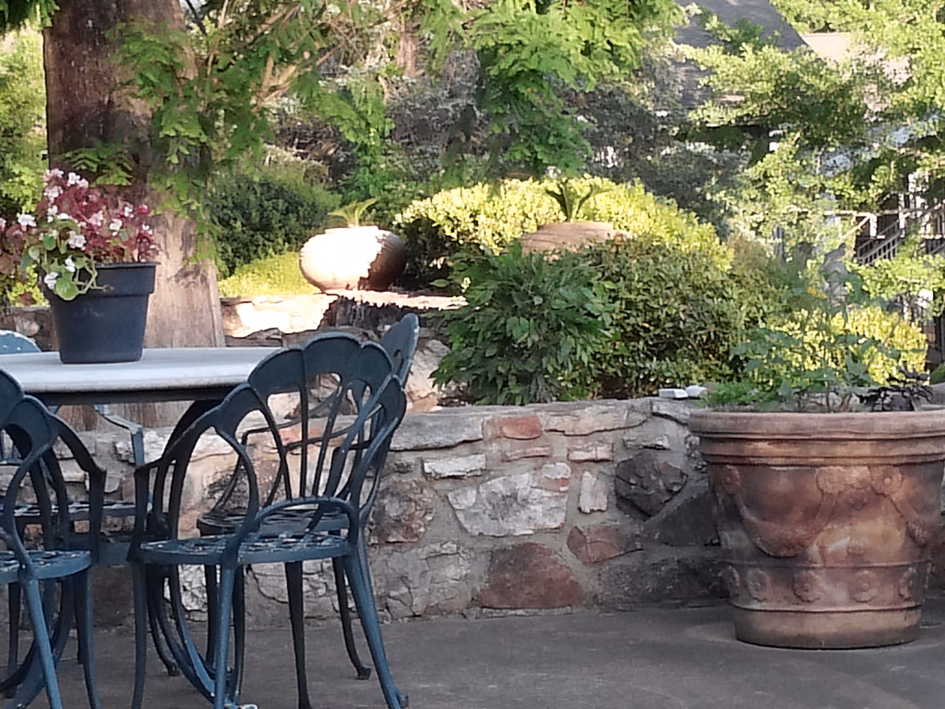 Outdoor patio area with metal chairs and a table, a potted plant on the table, large decorative pots, stone wall, and lush green bushes and trees in the background.