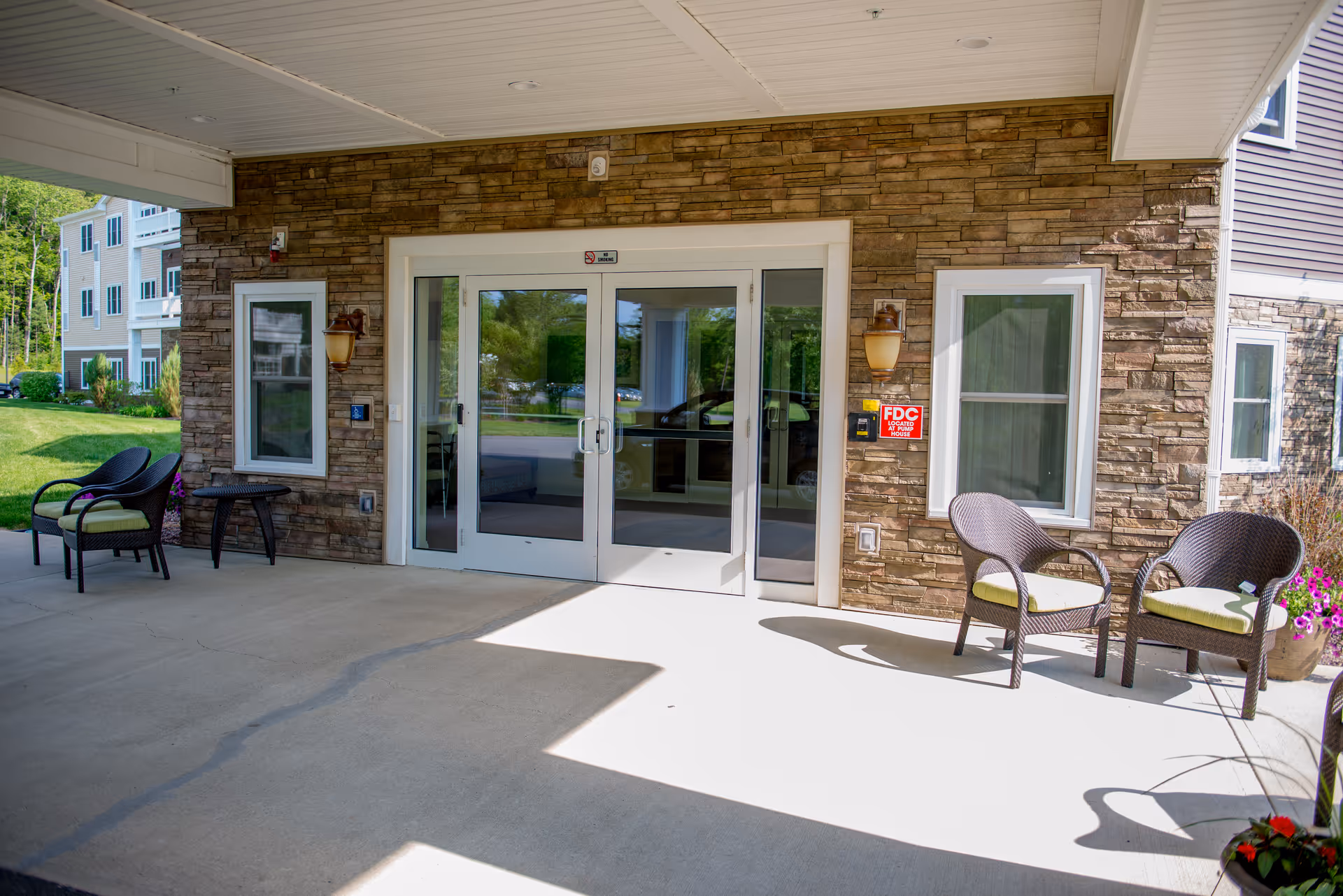 Covered entrance area of a building with stone facade walls, two glass double doors in the center, two windows on either side, outdoor chairs with cushions, a small table, and some potted plants. The area is well-lit with natural sunlight and has a concrete floor.