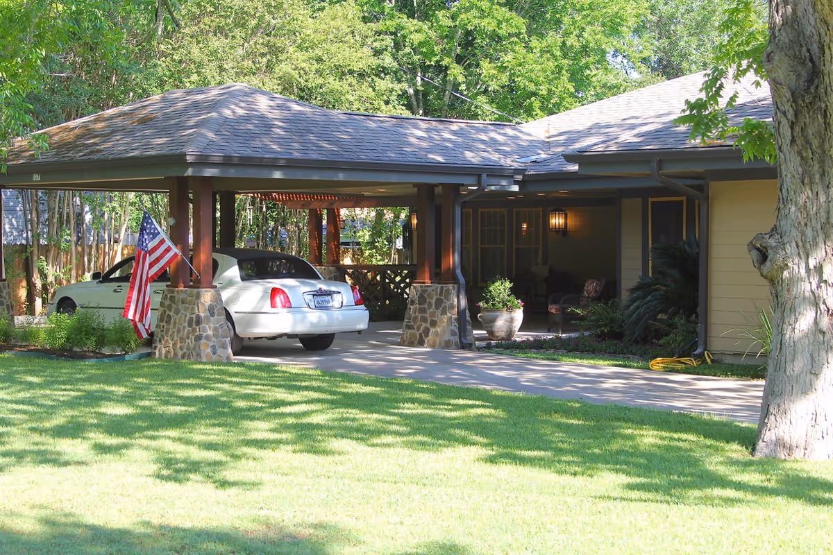 A covered carport attached to a house with a white car parked underneath. The carport has stone pillars and wooden beams. An American flag is mounted on one of the pillars. The house has a porch with outdoor seating and potted plants. The surrounding area includes a well-maintained lawn and trees.