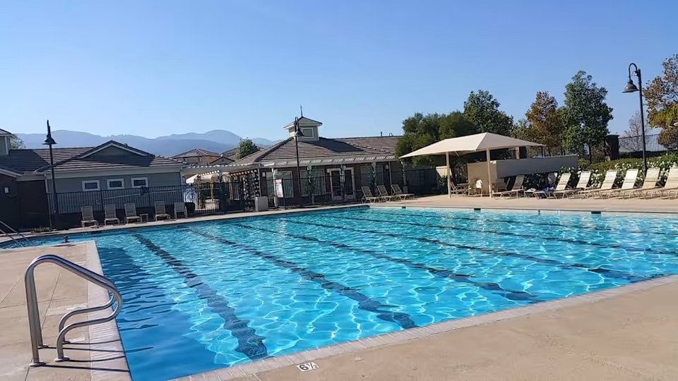 Outdoor swimming pool with lane markings, lounge chairs and a shaded seating area in front of a low building with mountains in the background.