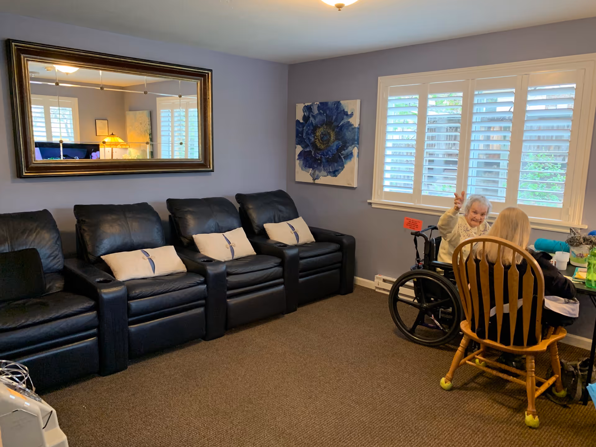 A cozy living room with four black leather recliners each with a white pillow, a large rectangular mirror on the wall, and a blue flower painting. Two women are seated near a window with white shutters; one woman is in a wheelchair waving, and the other is seated on a wooden chair with tennis balls on the legs.