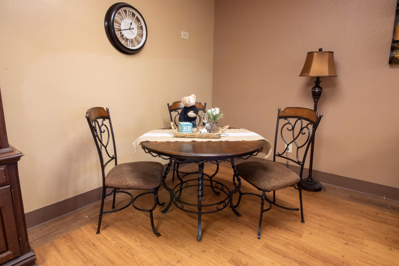 A small dining area with a round wooden table covered partially with a beige table runner. On the table, there is a teddy bear, a small sign that says 'BE', and a vase with white flowers. The table is surrounded by three metal chairs with cushioned seats. A wall clock is mounted on the beige wall above the table, and a floor lamp with a brown lampshade stands in the corner. The floor is wooden.