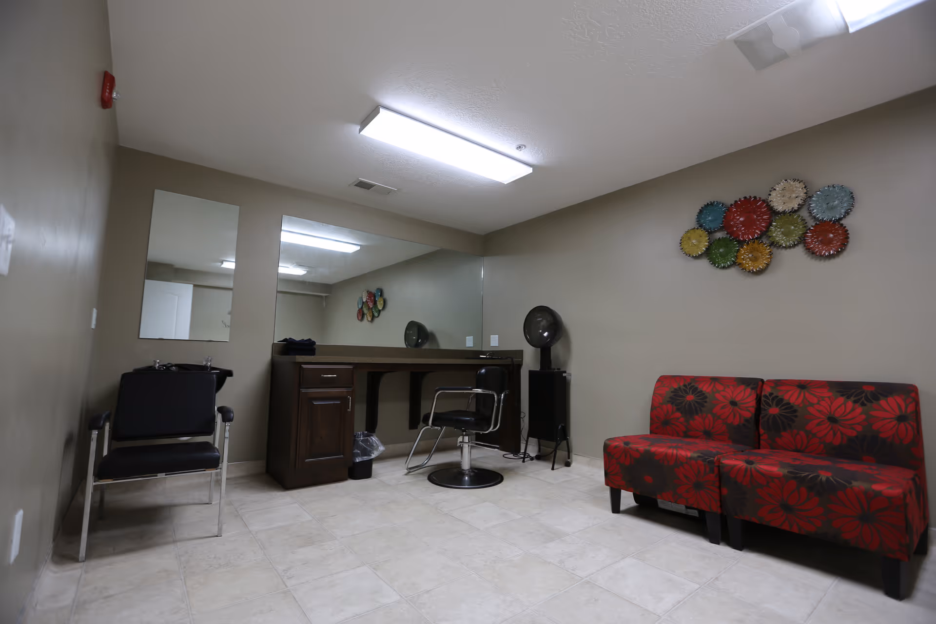 Interior room with beige walls and tiled floor featuring a black salon chair, a styling station with a large mirror, a black hair dryer on a stand, and a red floral-patterned bench. Colorful circular wall decorations are mounted above the bench.