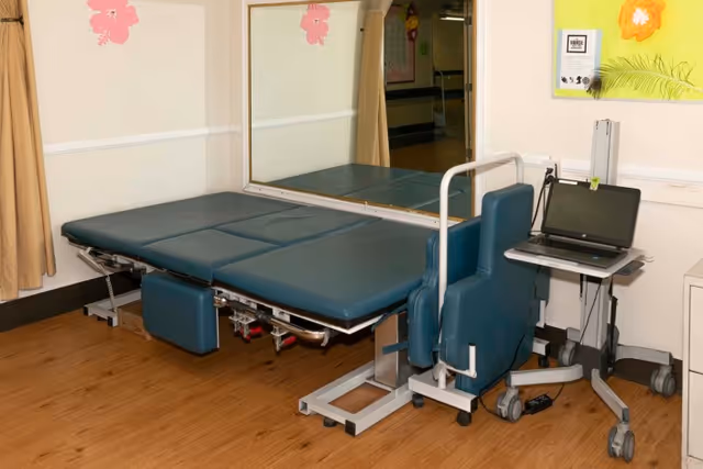 A medical examination or treatment room with a padded examination table, a blue padded chair with armrests on wheels, a laptop on a mobile stand, a large mirror on the wall, and light-colored walls with a yellow bulletin board decorated with a flower.