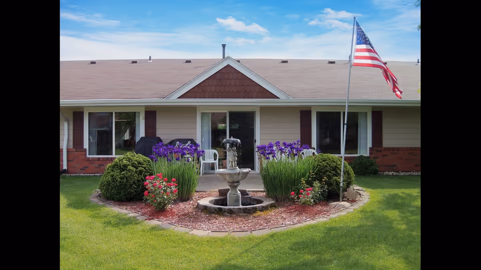 Front view of a single-story building with beige siding and a brown shingled roof. In front of the building is a circular garden with a stone fountain in the center, surrounded by purple flowers, green bushes, and red flowers. An American flag on a flagpole is positioned to the right of the garden. The building has large windows and a sliding glass door leading to a small patio with white plastic chairs.