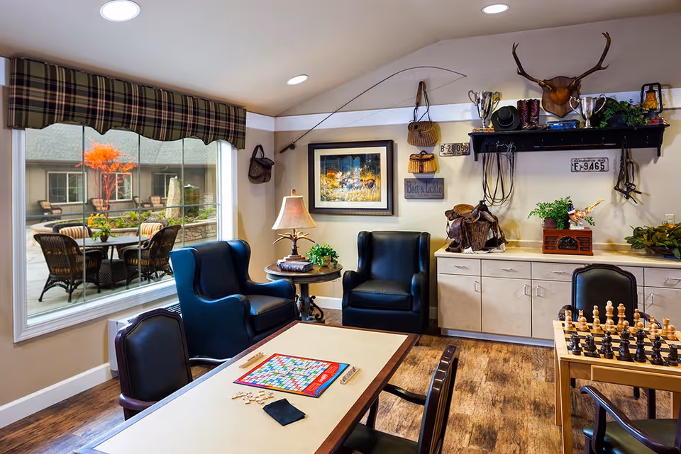 A cozy common area with two black leather armchairs, a table with a Scrabble board game, and another table with a chess set. The room has a large window with a plaid valance showing an outdoor patio with chairs and a small tree with orange leaves. The walls are decorated with a mounted deer head, trophies, framed artwork, and various rustic items including baskets and a fishing rod. There is a cabinet with plants and decorative items underneath a black shelf.