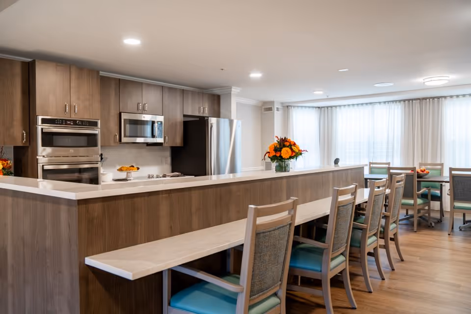 A modern kitchen and dining area in a senior living facility featuring wooden cabinetry, stainless steel appliances including an oven, microwave, and refrigerator, a long counter with a vase of orange flowers, and several wooden chairs with teal cushions arranged along the counter and around tables near large windows with sheer white curtains.