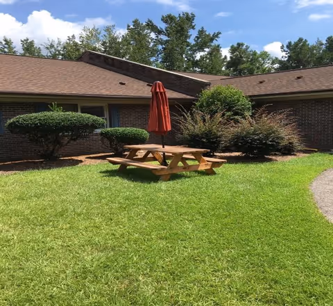 Outdoor area with a wooden picnic table and a closed red umbrella in the center, surrounded by green grass, bushes, and a brick building under a partly cloudy blue sky.
