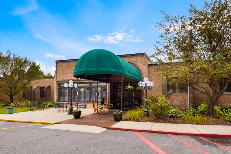 Exterior view of a brick building with a green canopy over the entrance. There are outdoor chairs and tables on the patio, surrounded by trees and plants under a partly cloudy blue sky.