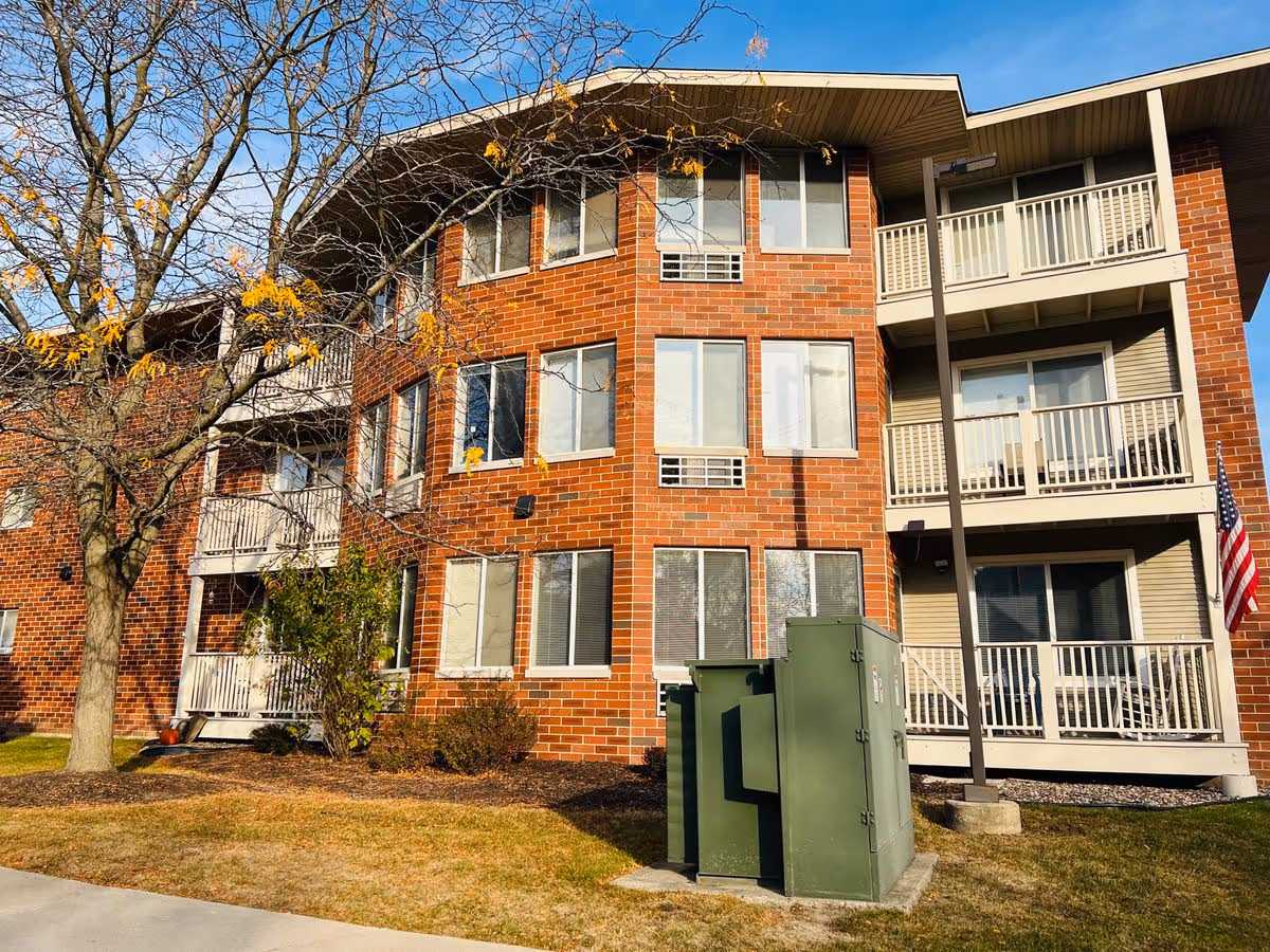 Three-story brick senior living building with balconies, a leafless tree in front, and green utility boxes on the lawn.