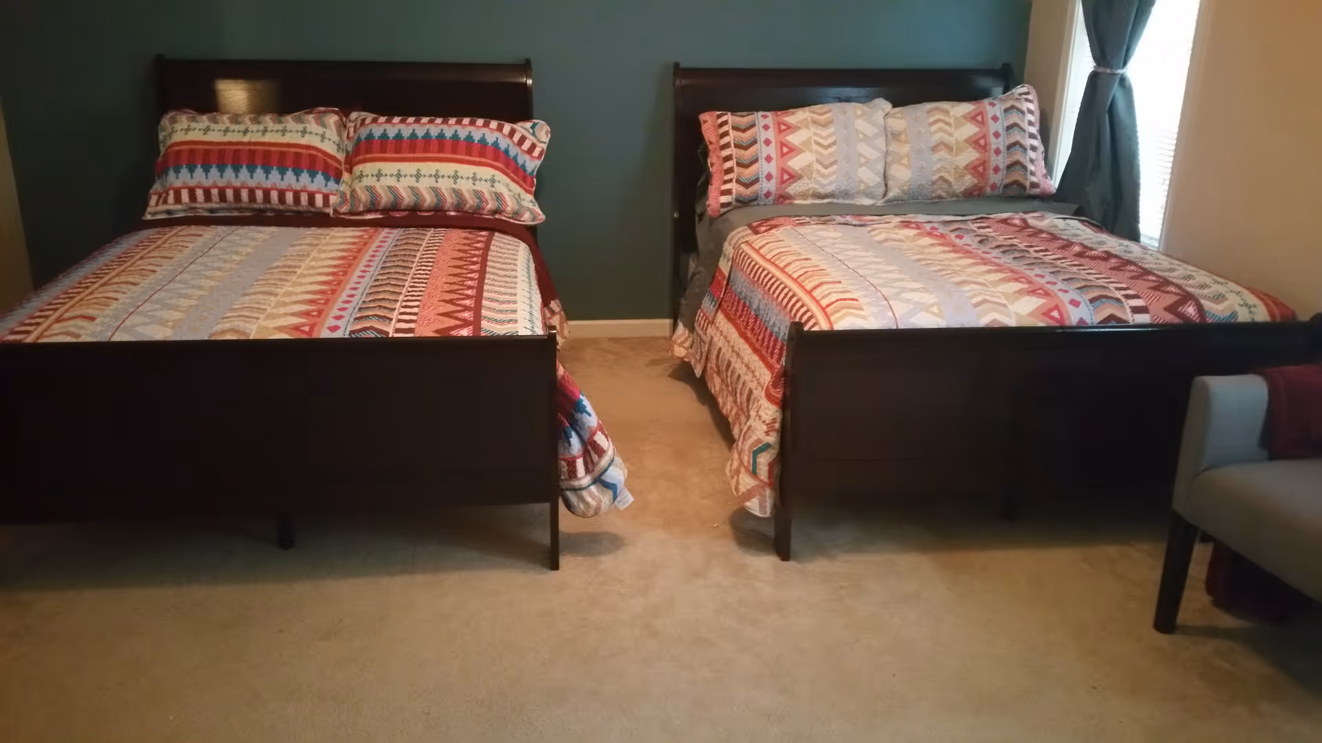 A bedroom with two dark wooden beds side by side, each made with colorful patterned bedspreads and matching pillows. There is a window with curtains on the right side and a gray armchair partially visible in the corner.