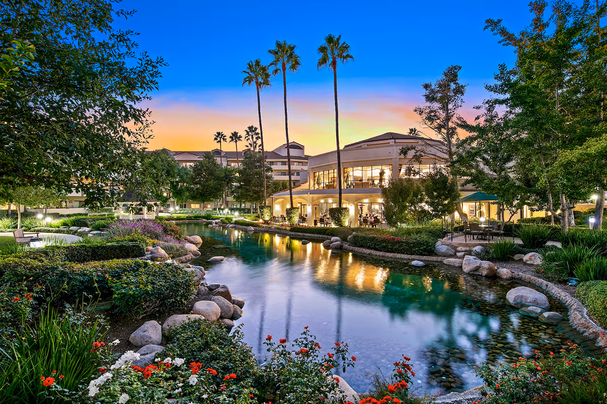 A serene outdoor garden area at sunset with a clear pond surrounded by rocks, lush greenery, colorful flowers, and tall palm trees. In the background, a large building with illuminated windows and outdoor seating is visible.