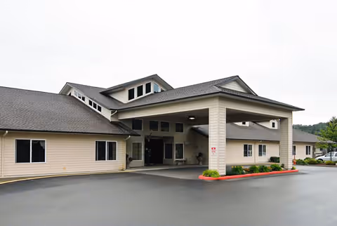 Exterior view of the entrance to a single-story senior living facility building with beige siding, a covered drop-off area supported by columns, and a paved driveway in front.
