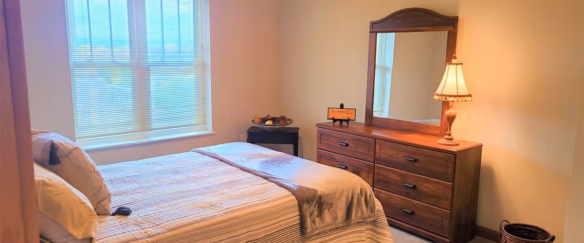 A bedroom with a neatly made bed featuring striped bedding and pillows, a wooden dresser with a large mirror, a table lamp, a small basket on the floor, and a window with blinds letting in natural light.
