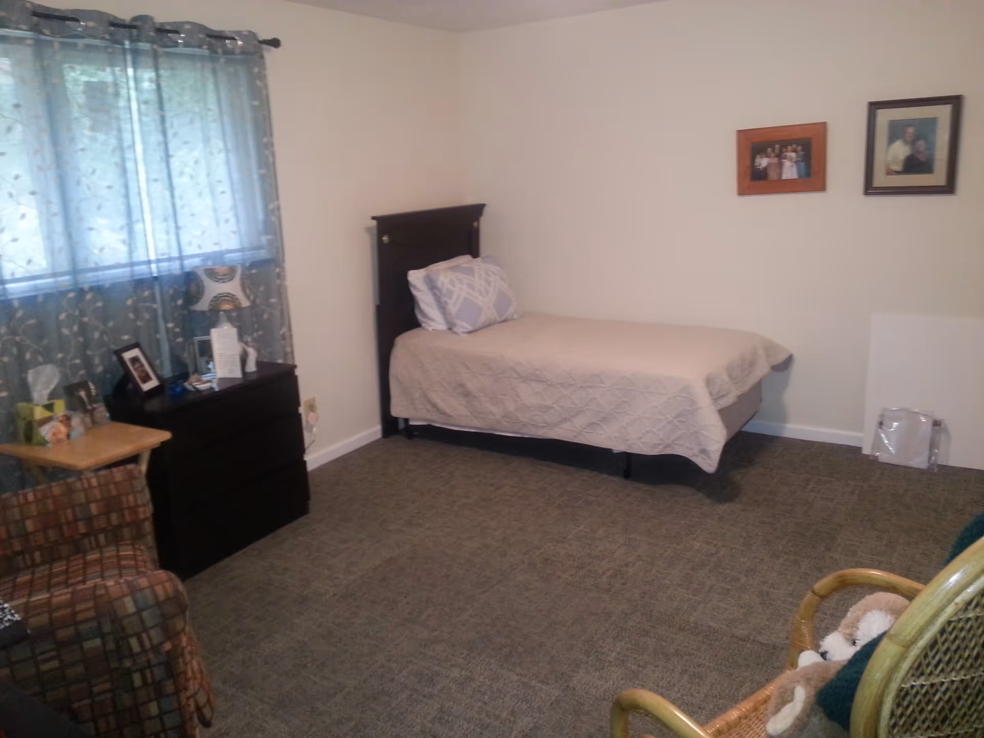A simple bedroom with a single bed covered in a beige quilt and a pillow, a dark wooden dresser with a lamp and framed photos on top, a window with sheer patterned curtains, a small wooden side table with tissues, a patterned armchair, and a wicker chair with a stuffed animal. Two framed pictures hang on the wall above the bed.