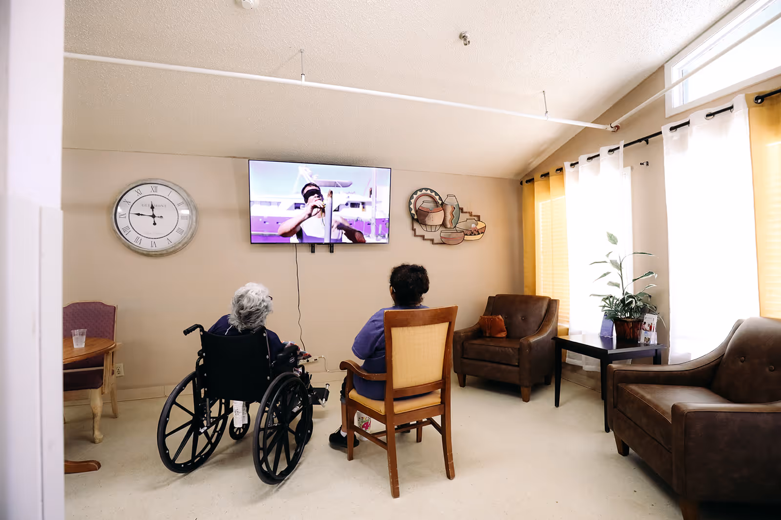 Two elderly individuals, one in a wheelchair and the other seated on a wooden chair, watching a television mounted on the wall in a well-lit room with large windows covered by white and yellow curtains. The room has brown armchairs, a side table with a plant, a wall clock, and decorative wall art.