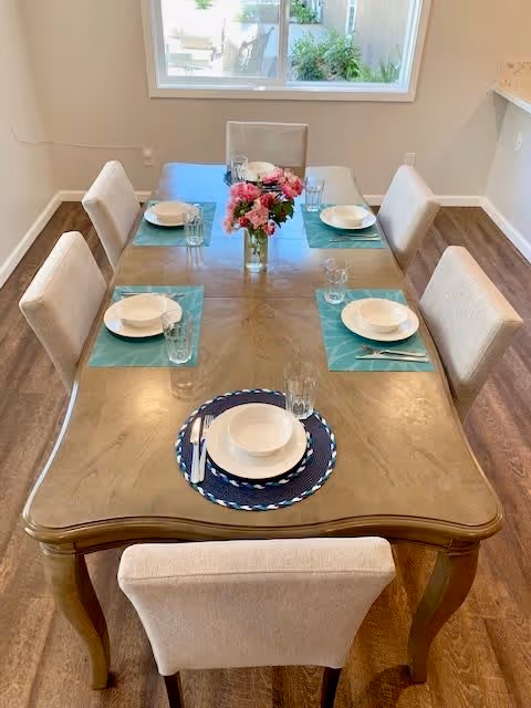 A dining room with a wooden table set for six people. Each place setting includes a white bowl, plate, glass, and silverware on blue or teal placemats. A vase with pink flowers is centered on the table. The room has wooden flooring and a window with a view of outdoor greenery.