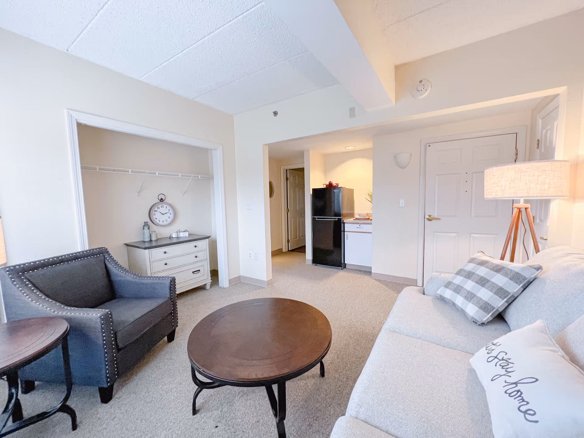 A cozy living room area in a senior living facility featuring a light-colored sofa with decorative pillows, a round wooden coffee table, a dark upholstered armchair, a small side table, and a floor lamp. In the background, there is an open closet with a white dresser and a wall clock, and a kitchenette area with a black refrigerator and white cabinets near the entrance door.