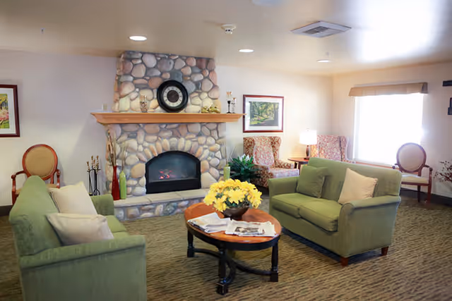 A cozy living room with two green sofas facing each other around a wooden coffee table with a yellow flower arrangement. The room features a stone fireplace with a clock and decorative items on the mantel. There are two armchairs with floral upholstery near a window with a beige valance, and framed artwork on the walls.