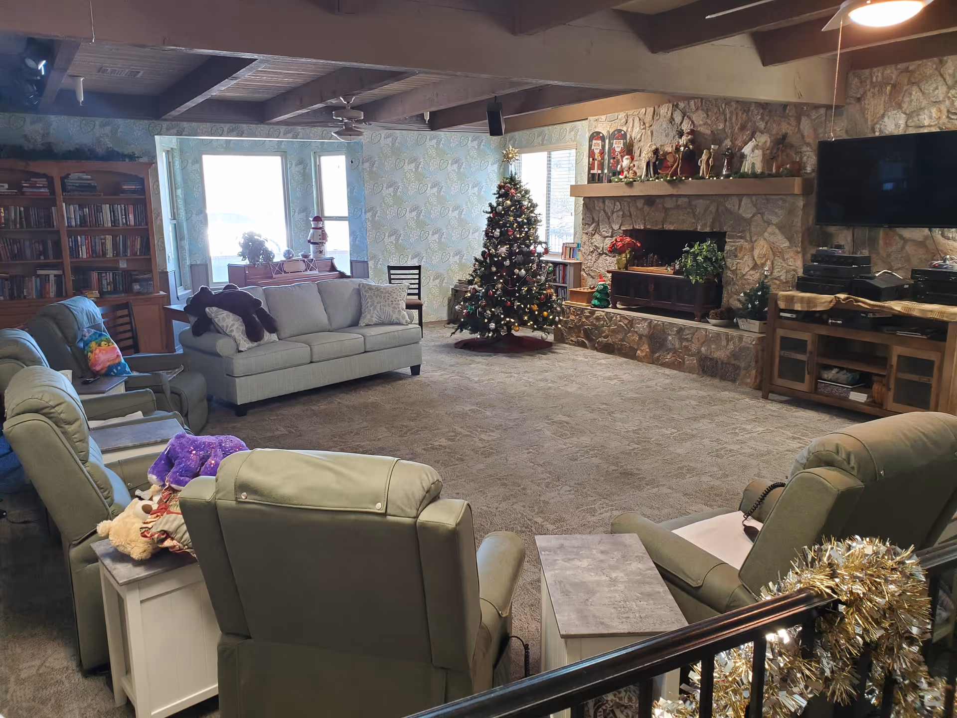 Spacious communal living room with sofas and recliners arranged around a stone fireplace and a decorated Christmas tree.