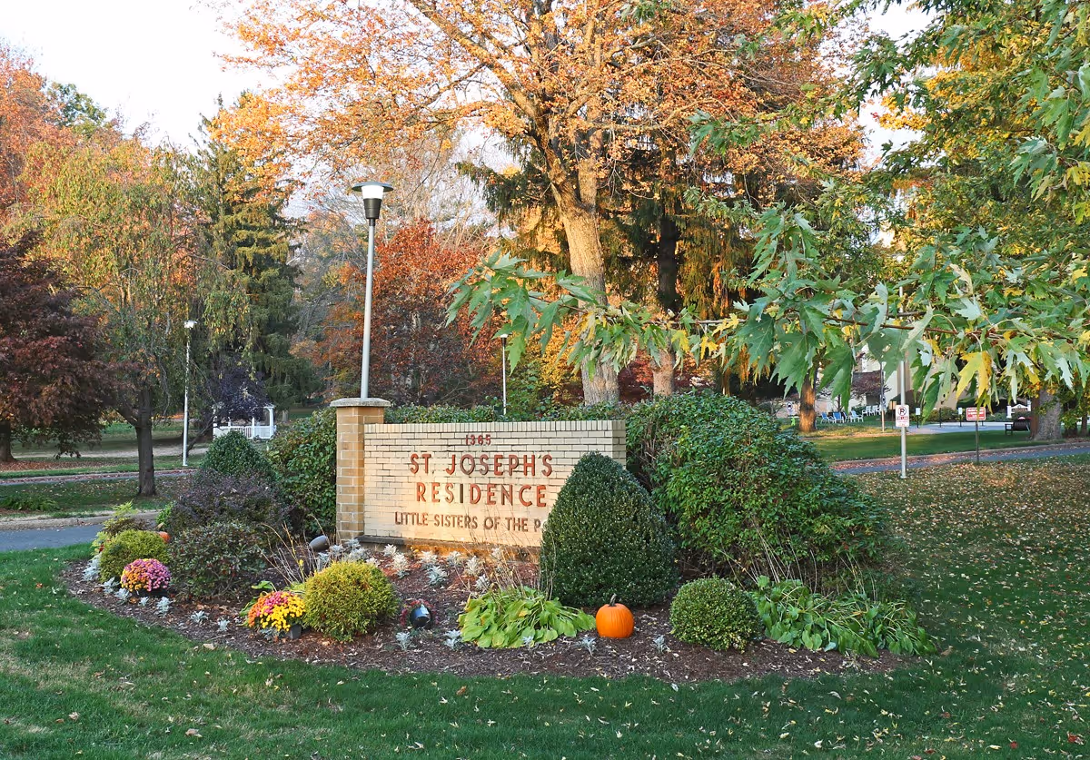 Outdoor view of a landscaped area with a brick sign that reads '1365 St. Joseph's Residence Little Sisters of the Poor' surrounded by bushes, flowers, and a pumpkin, with trees showing autumn foliage in the background.