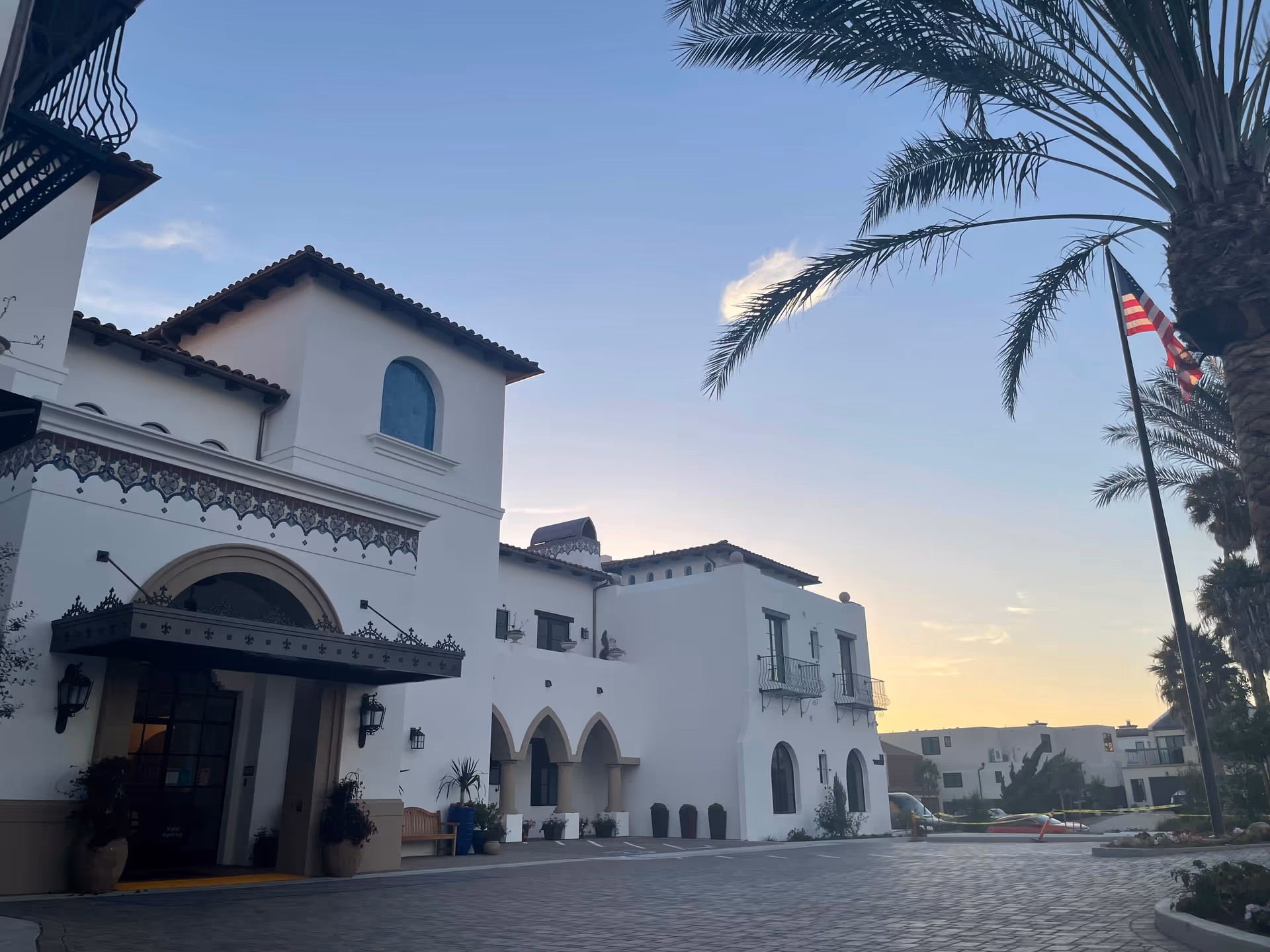 Exterior view of a white building with Spanish-style architecture at sunset, featuring arched doorways, decorative tiles, palm trees, and an American flag on a flagpole.
