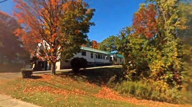Exterior view of a single-story building with a green roof surrounded by trees with autumn foliage and a grassy area with fallen leaves.