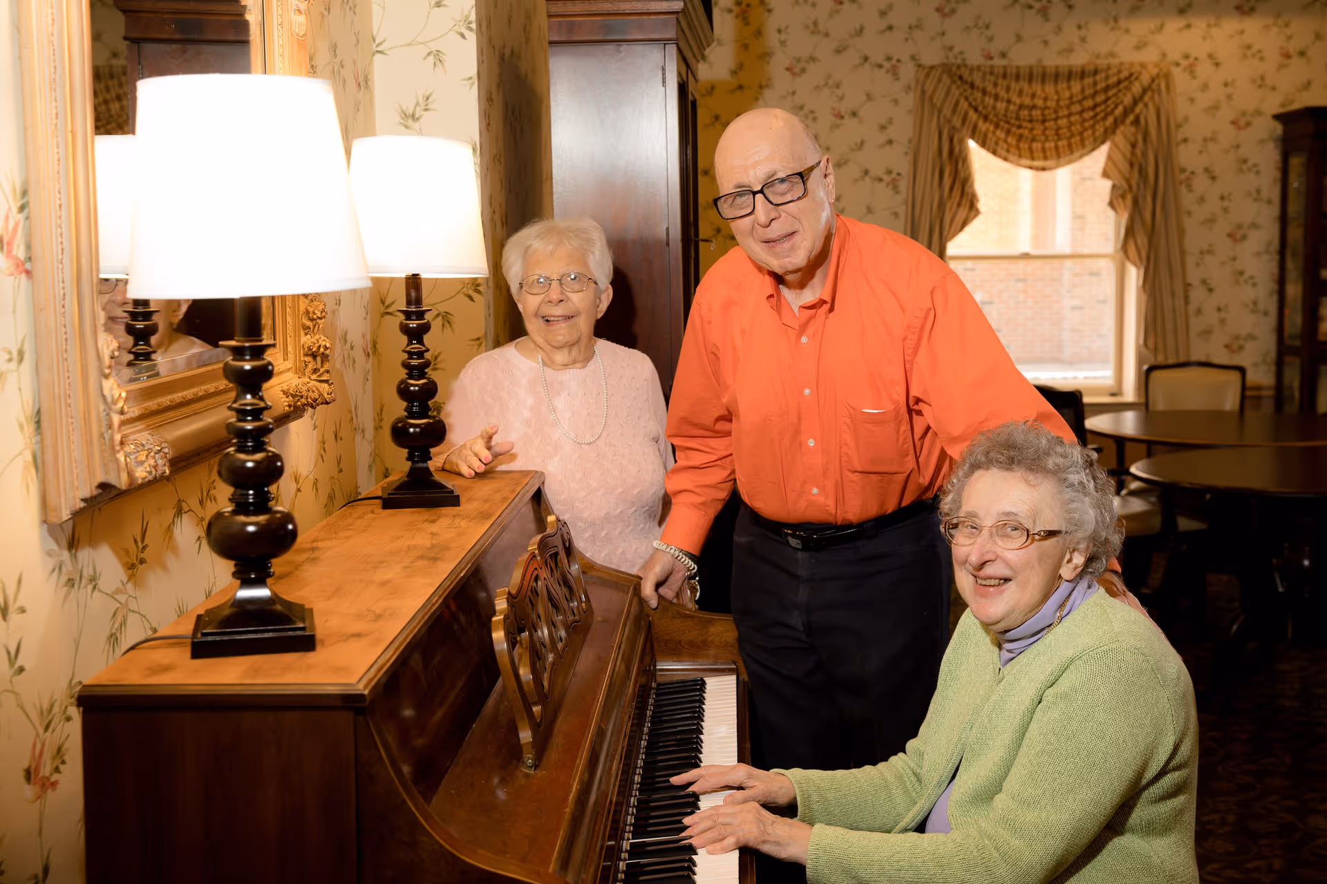 Three elderly people in a warmly lit room with floral wallpaper. One woman is seated at a wooden piano playing, while a man in an orange shirt and another woman in a light pink sweater stand nearby, smiling. The room has a large mirror, two table lamps on the piano, and a window with draped curtains in the background.