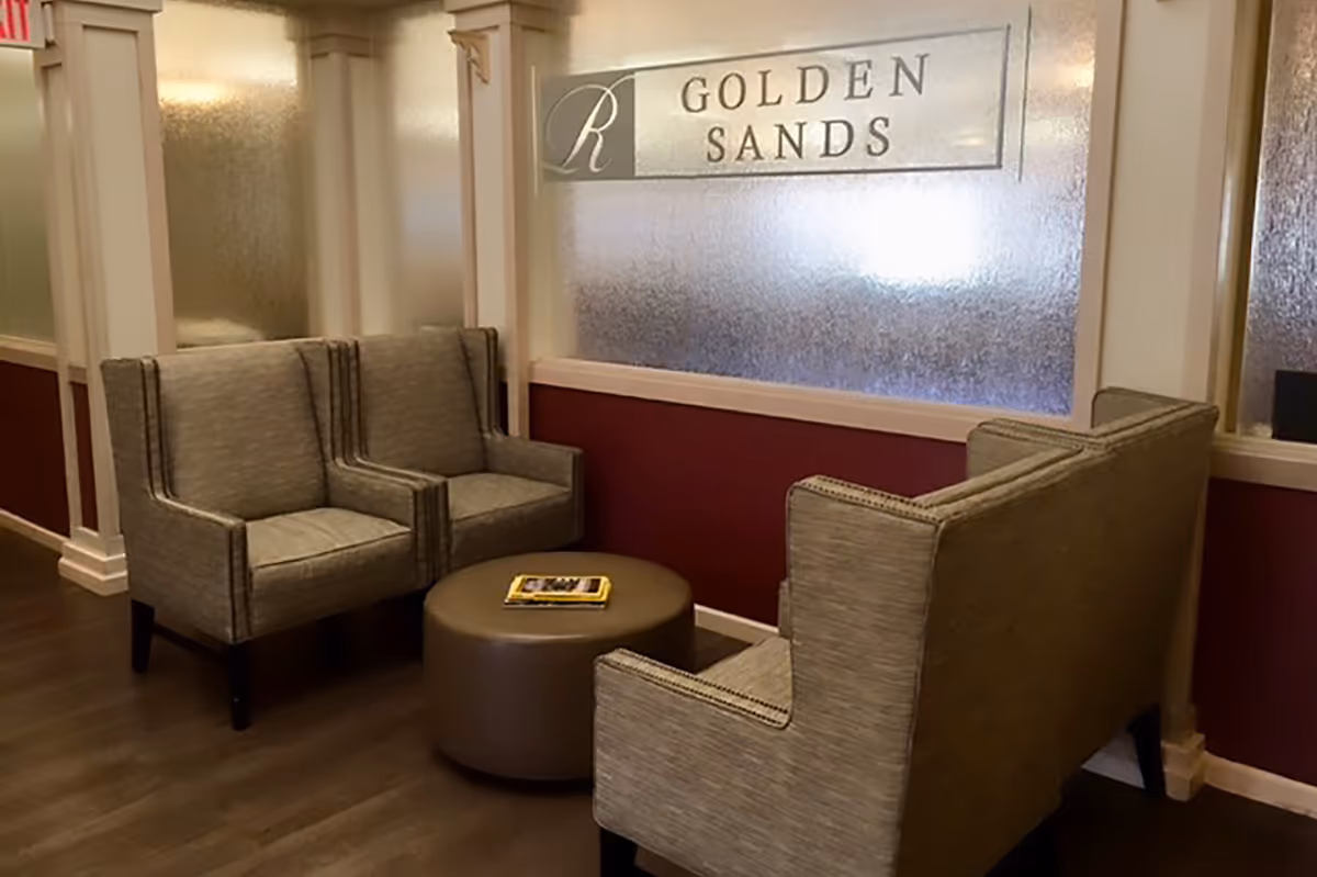 A seating area in a facility with three gray upholstered armchairs arranged around a round brown ottoman with a magazine on top. Behind the chairs is a frosted glass panel with the text 'Golden Sands' and a decorative 'R' logo. The walls are painted in a combination of white and dark red, and the floor is wooden.