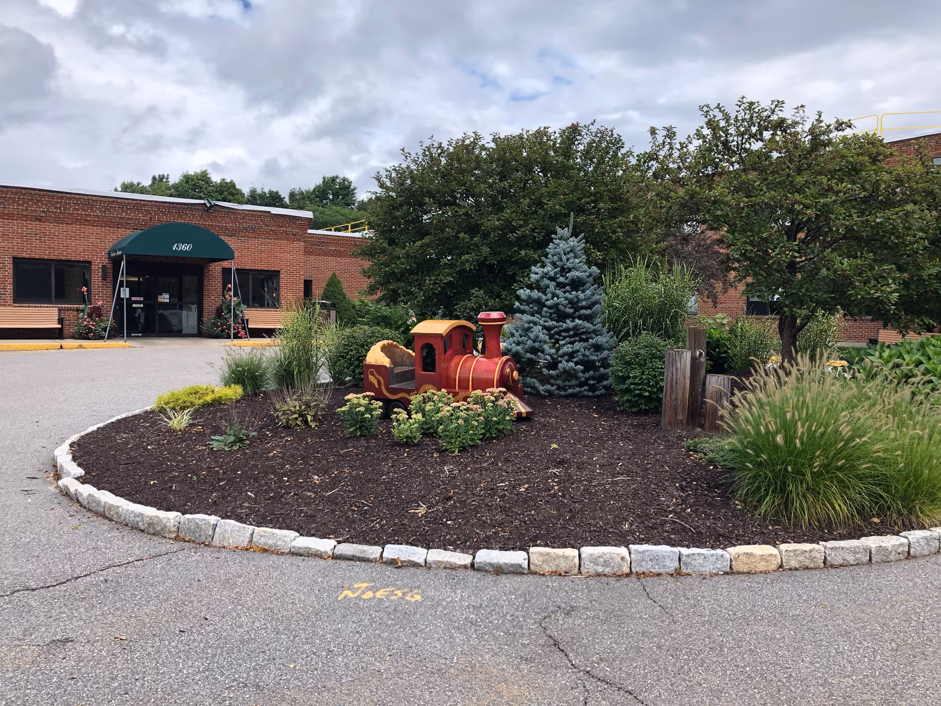 Exterior view of Havencare at Valerie Manor showing a brick building with a green awning over the entrance. In front of the building is a landscaped circular garden bed with various plants, shrubs, and a small decorative red and yellow train. The sky is cloudy.