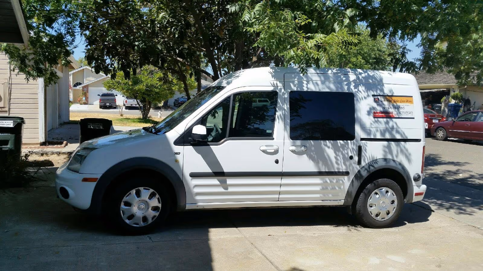 A white van parked on a driveway next to a house with trees casting shadows on the vehicle and surrounding area. The van has a sign on the rear side panel with contact information. There are other cars and houses visible in the background.