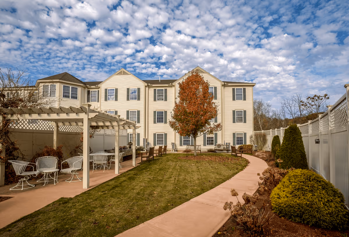Outdoor garden area at Benchmark at Billerica Crossings featuring a paved walkway, green lawn, a tree with autumn leaves in the center, white pergola with white metal chairs and tables on the left, benches along the walkway, and a three-story beige building with multiple windows in the background under a partly cloudy sky.