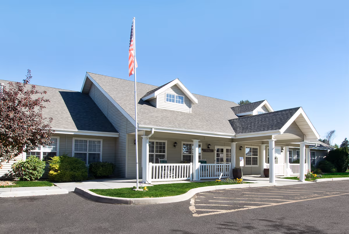 Exterior view of a single-story senior living facility building with a gray roof, beige siding, white trim, and a covered entrance porch. There is an American flag on a flagpole in front of the building, surrounded by neatly trimmed bushes and a clear blue sky.