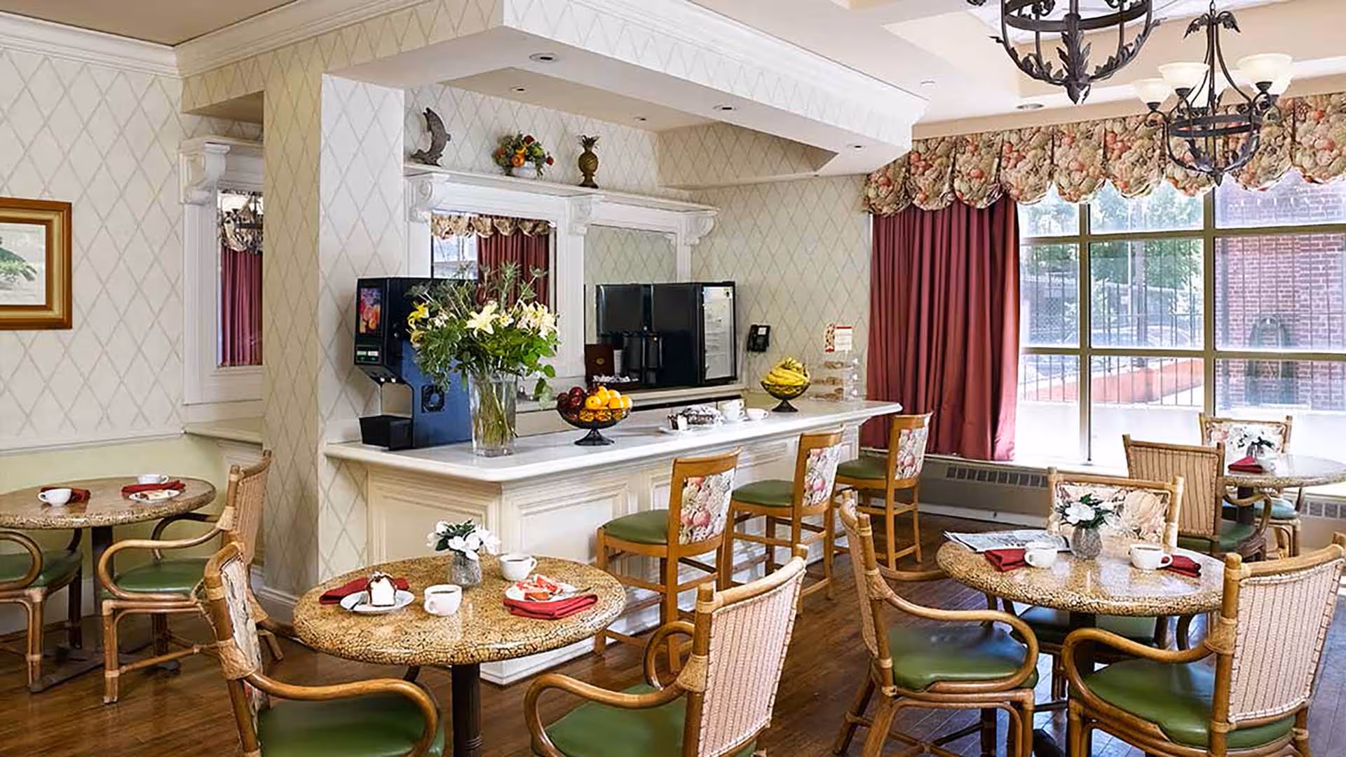A cozy dining area with round tables and wicker chairs with green cushions. A counter with bar stools featuring floral upholstery is set against a wall with a coffee machine, fruit bowls, and decorative items. Large windows with floral valances and red curtains allow natural light to fill the room.