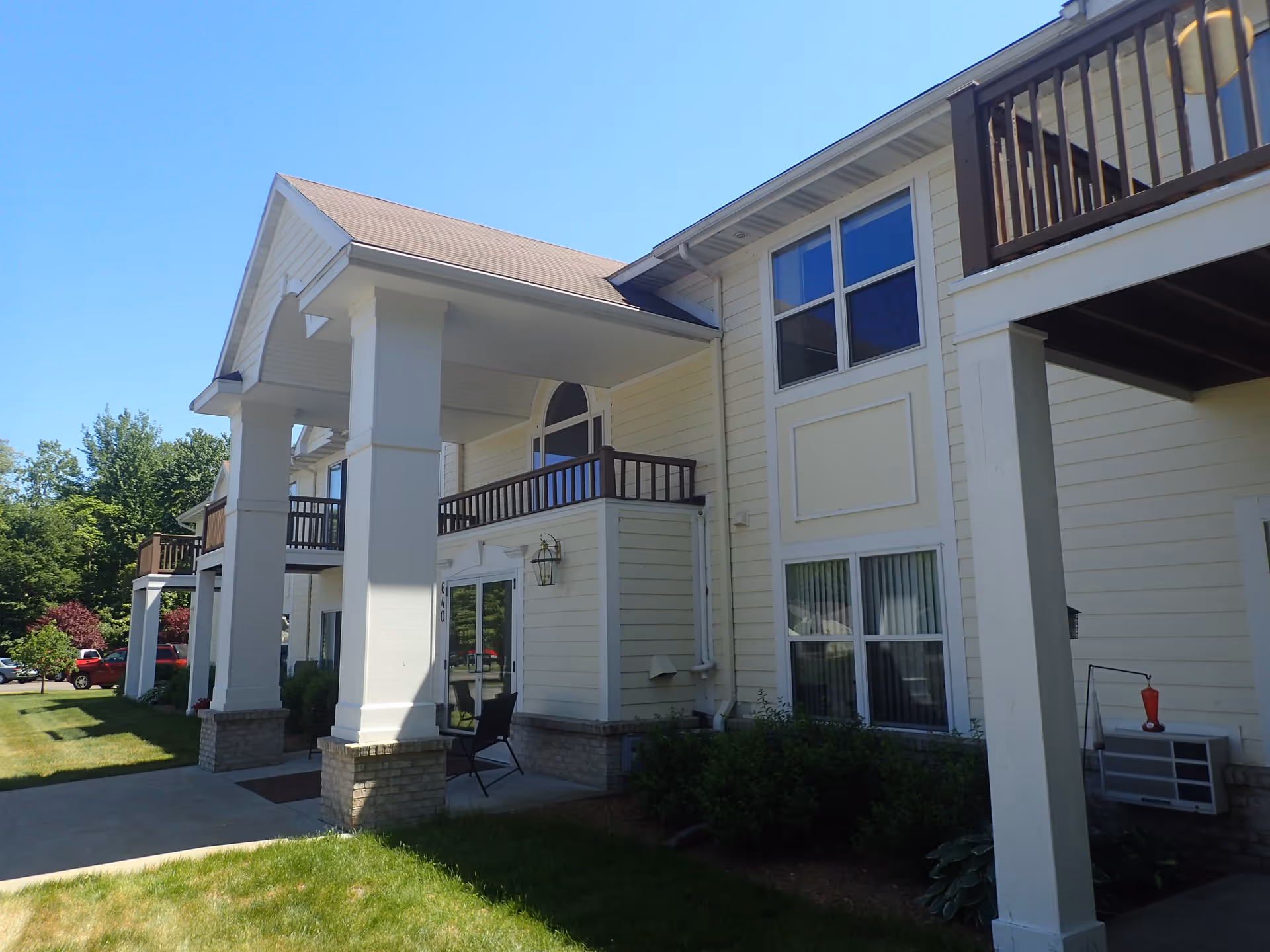 Exterior view of a two-story residential building with white siding, large columns supporting a covered entrance, balconies with dark railings, and a well-maintained lawn under a clear blue sky.