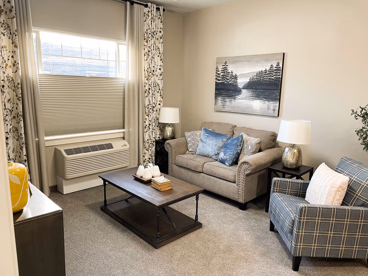 A cozy living room with a beige loveseat adorned with blue and patterned pillows, a plaid armchair with a white pillow, a dark wooden coffee table with decorative items, two side tables each with a lamp, floral curtains framing a window with a beige blind, and a landscape painting of a lake and trees on the wall.