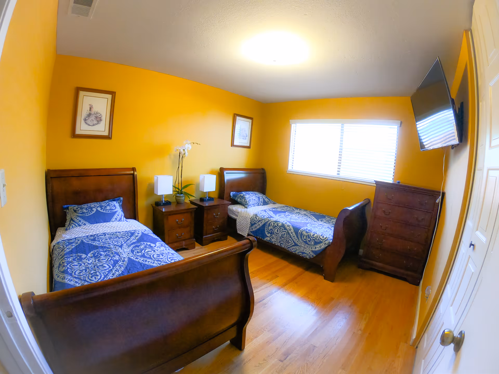 A bedroom with two single wooden beds with blue patterned bedding. Between the beds are two wooden nightstands with lamps and a white orchid plant. The walls are painted yellow, and there are two framed pictures hanging above the beds. A window with blinds is on the far wall, and a flat-screen TV is mounted on the right wall above a wooden dresser. The floor is wooden.