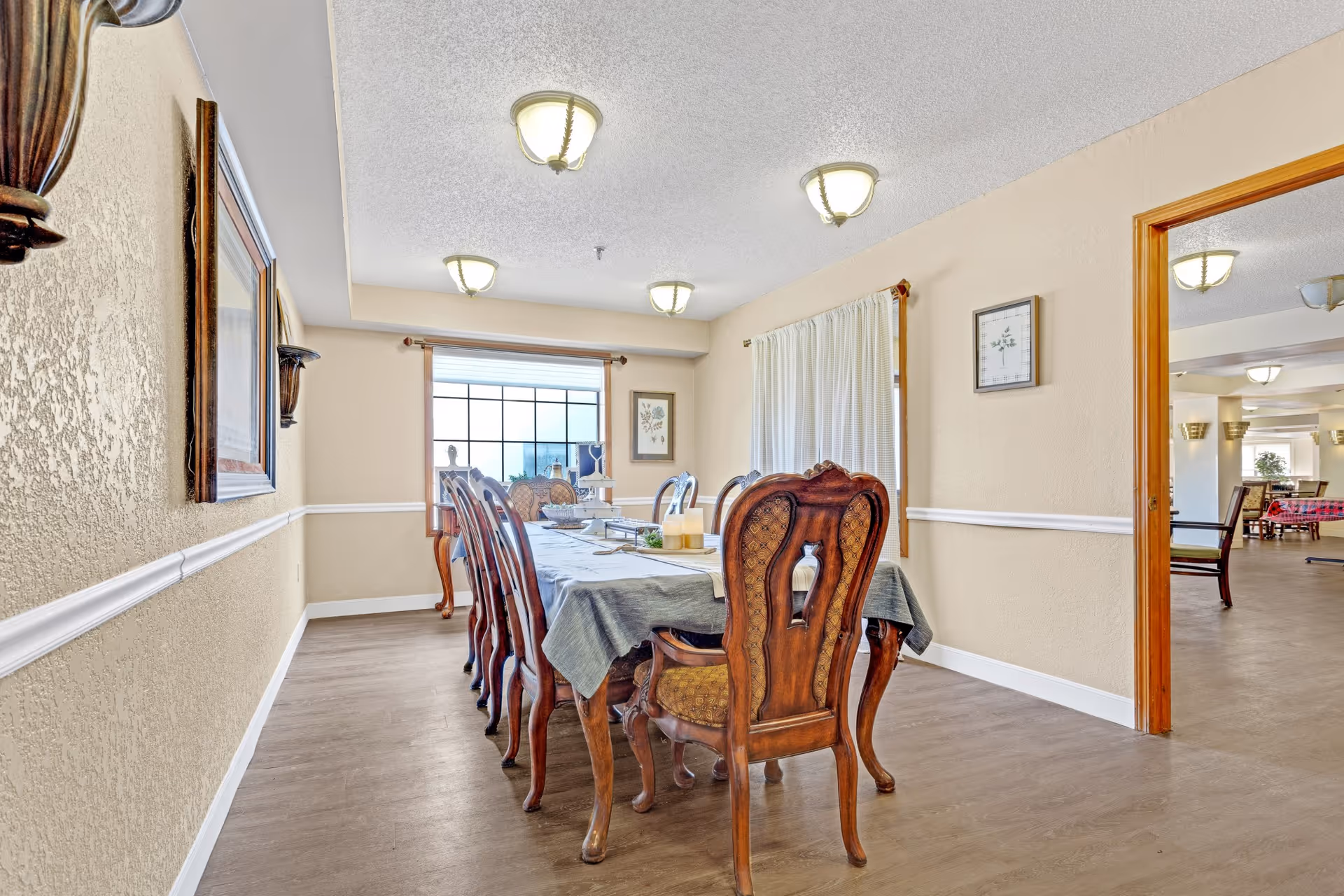 A dining room with a long wooden table covered with a gray tablecloth and surrounded by ornate wooden chairs. The room has beige walls with white trim, a large window with curtains, framed artwork on the walls, and ceiling lights. An open doorway leads to another room with more seating visible.