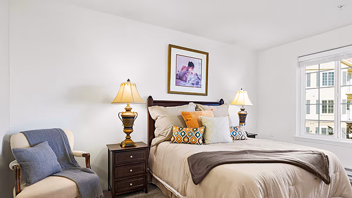 Bright bedroom with a made bed, two bedside lamps, a nightstand, an armchair with a throw, and a window showing neighboring buildings.