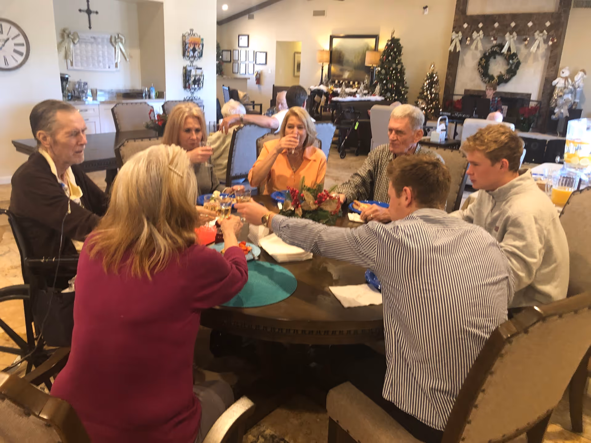 A group of elderly and middle-aged people sitting around a round dining table in a decorated room, raising their glasses in a toast. The room has Christmas decorations including a Christmas tree, wreaths, and garlands. There is a clock on the wall and a cross hanging above a calendar in the background.