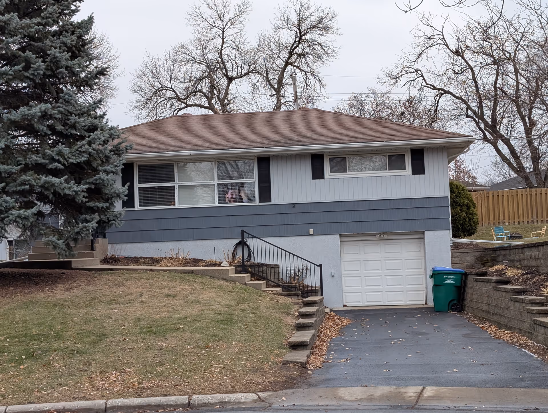 A single-story residential building with a sloped roof, gray and white exterior, a garage with a white door, and a driveway. There is a large evergreen tree on the left side and leafless trees in the background. The lawn is mostly brown with some patches of green. A green trash bin is placed near the garage door, and there are two chairs and a small table on a raised grassy area to the right.