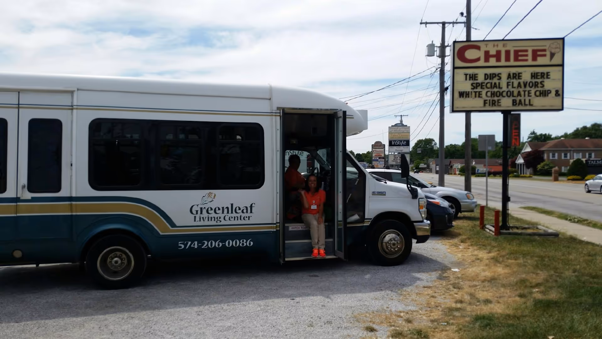 A Greenleaf Living Center shuttle bus parked on the side of a road with its door open. Two people are sitting inside the bus near the entrance. In the background, there is a street with cars and a sign for The Chief advertising special ice cream dip flavors.