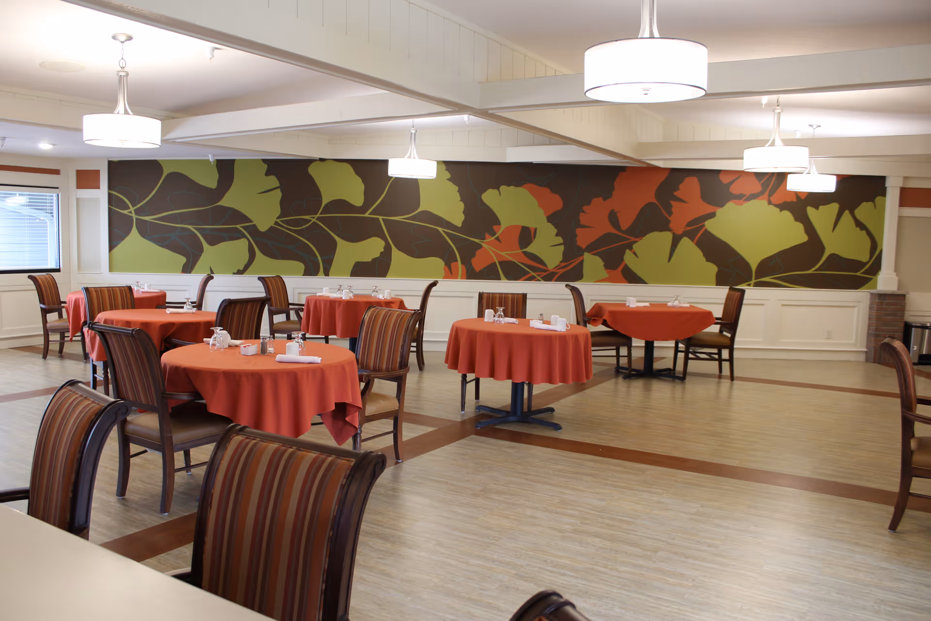 Dining room with round tables covered in red tablecloths, each set with glasses, napkins, and condiments. The room has striped wooden chairs and a large decorative wall panel featuring a leaf pattern in green, brown, and orange colors. Ceiling lights hang above the tables, and there is a window on the left side.